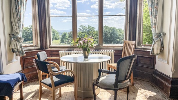 Quarto com uma mesa redonda, quatro cadeiras, um radiador e uma janela com vista para vegetação e céu nublado.
