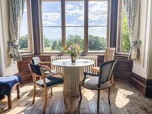 A room with a round table, four chairs, a radiator, and a window with a view of greenery and a cloudy sky. - Nethercott House (Winkleigh)