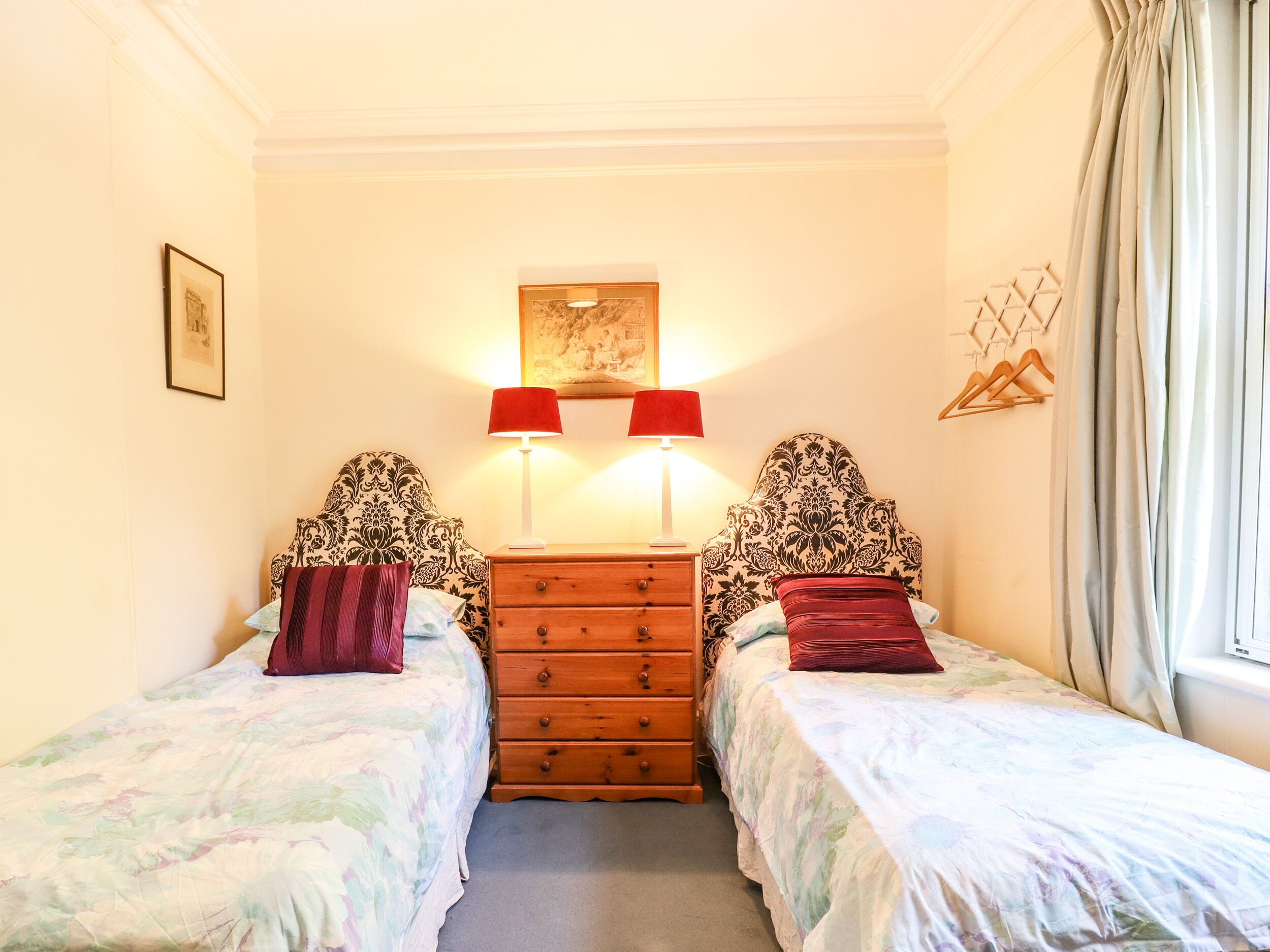 Une chambre avec deux lits simples, une commode en bois, deux lampes rouges et une œuvre d’art accrochée au mur.