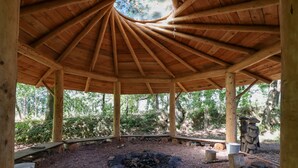 Um pavilhão de madeira com teto circular, uma fogueira no centro e uma estrutura de pedra à direita.