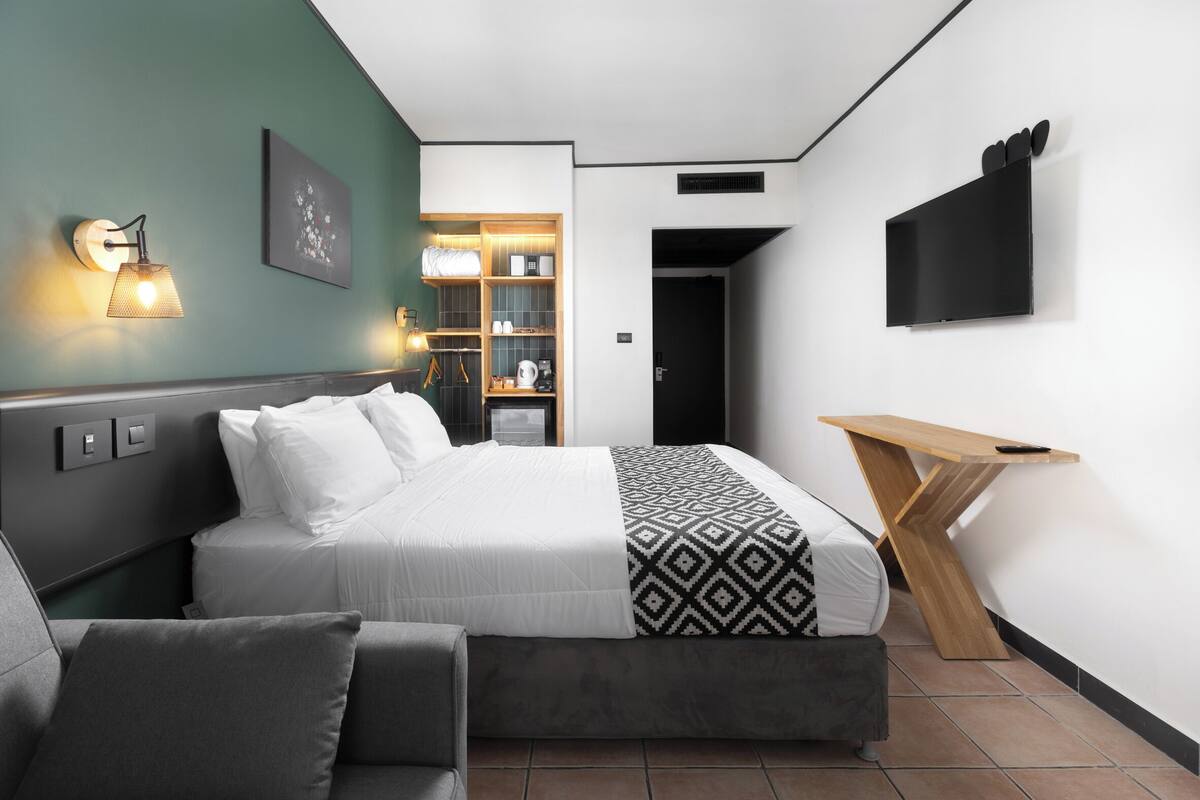A modern hotel room with a bed, a sofa, a TV mounted on the wall, and a wooden side table.