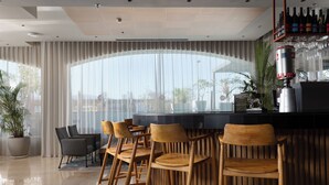 Uma área de bar moderna com banquetas de madeira, uma janela grande com cortinas leves e um balcão com uma máquina de café.