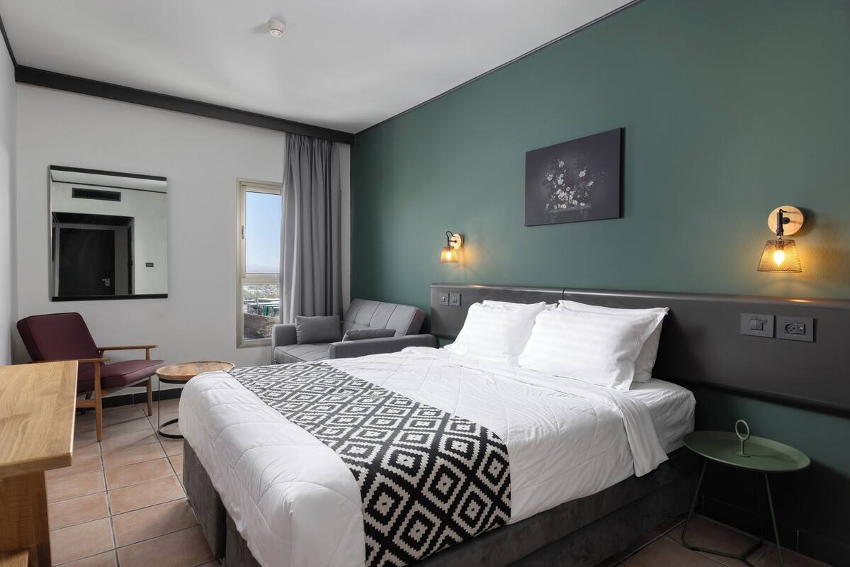 A hotel room with a bed, a desk, a chair, a small table, a sofa, and a view of the city.