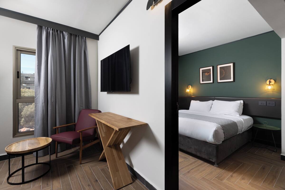 A modern hotel room with a wooden desk, a red chair, a bed with white bedding, and two wall-mounted picture frames.