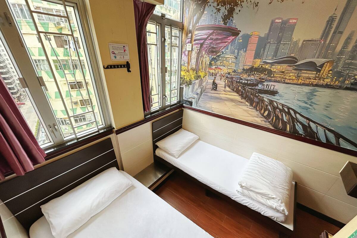 A hotel room with two single beds, a large window with a cityscape mural, and a view of the city.