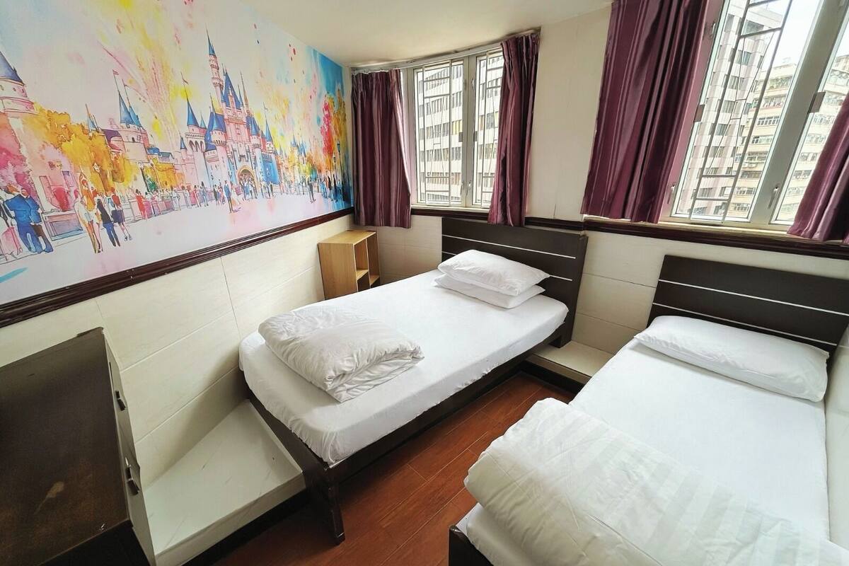 A hotel room with a large mural of a castle, a single bed with white bedding, a wooden nightstand, and a window with purple curtains.