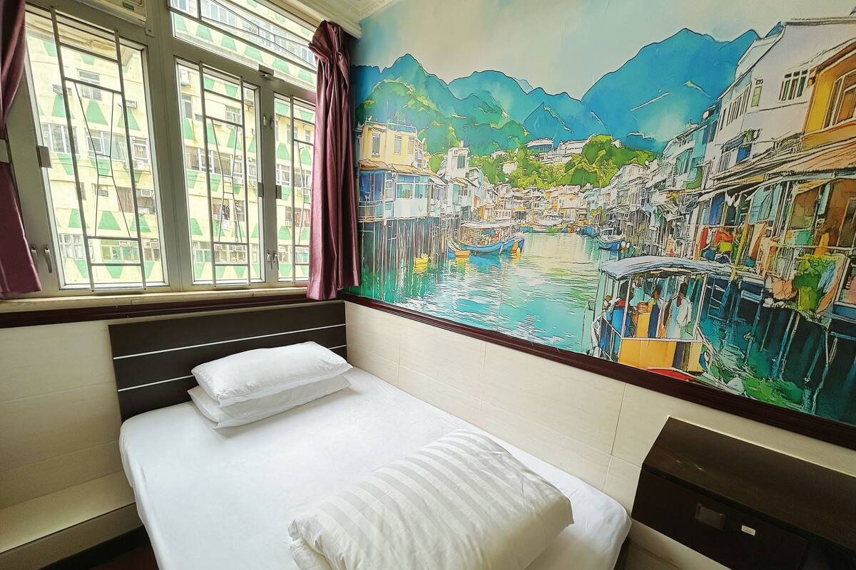 A hotel room with a bed, a window with curtains, and a mural of a riverside town.