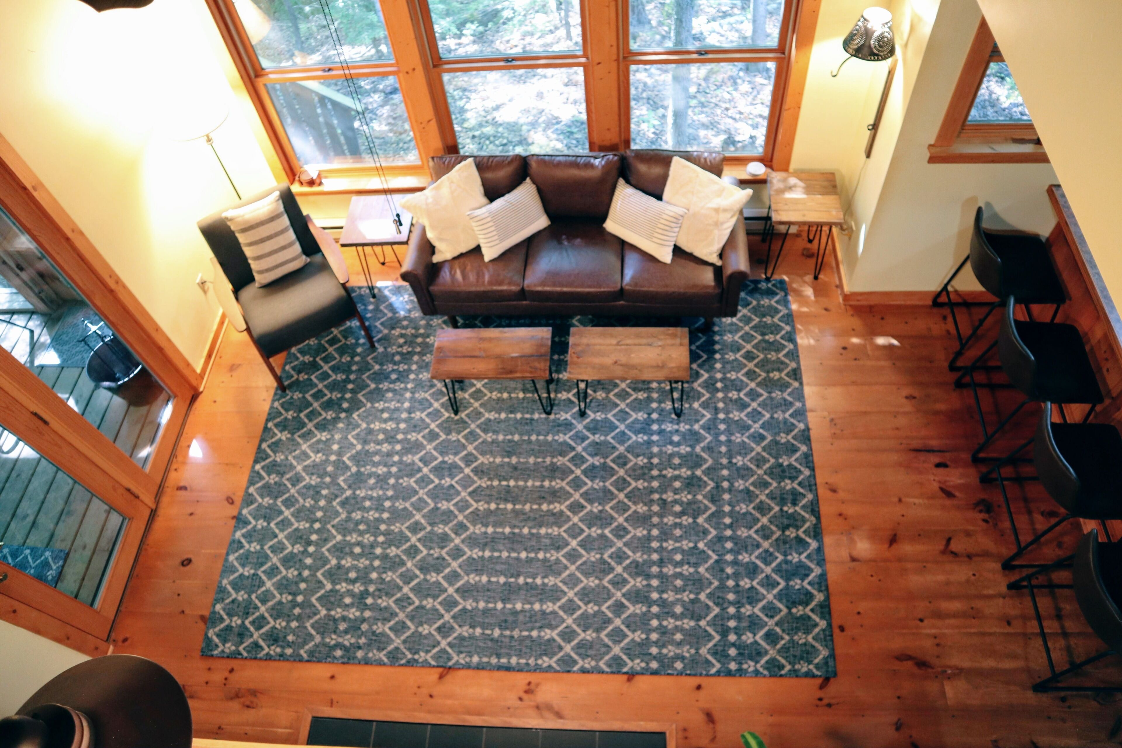 Una sala de estar acogedora con un sofá de cuero marrón, un tapete estampado, piso de madera y ventanas amplias con vistas a árboles.
