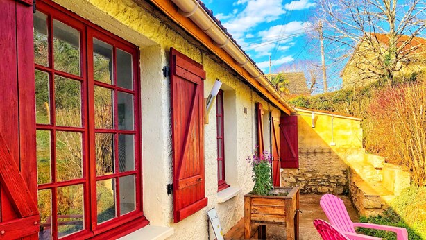 Una casa rústica con contraventanas rojas, un banco de madera y una silla rosa en el exterior.