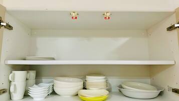Cozinha com armários brancos, bancada branca e prateleira branca. Há pilhas de pratos, tigelas e xícaras brancos, com uma tigela amarela na prateleira.