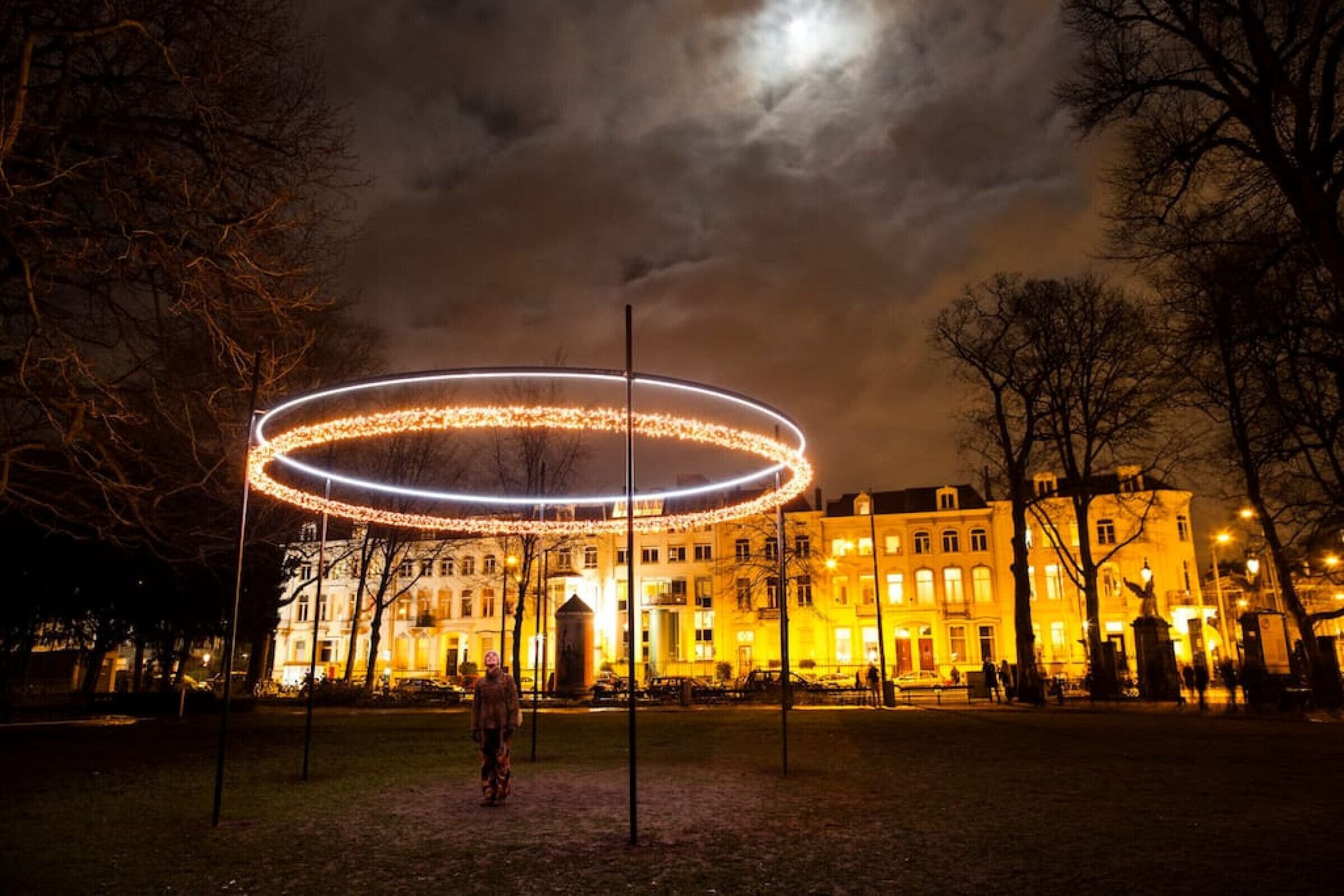 Une vue nocturne d’un parc, avec une installation lumineuse circulaire et un bâtiment en arrière-plan.