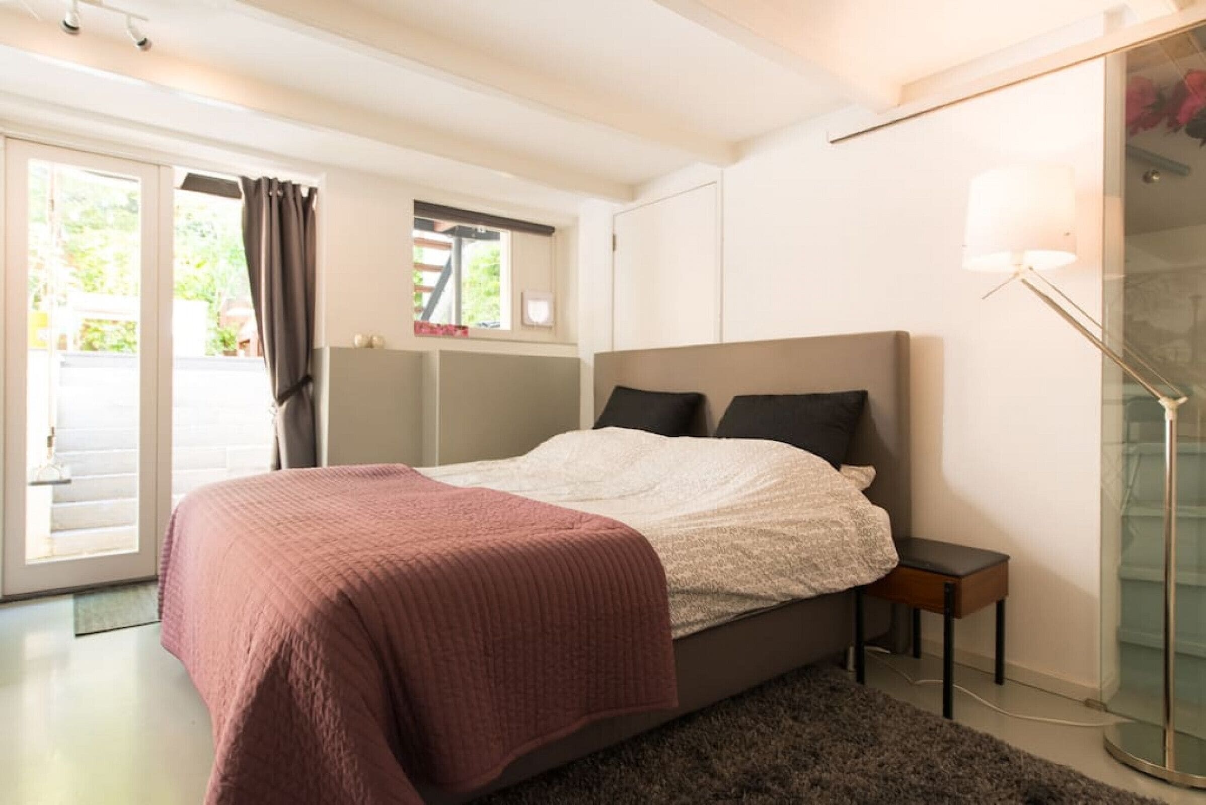 Une chambre à coucher comprenant un lit, une table de chevet, une lampe et une porte-fenêtre coulissante donnant sur un espace extérieur.