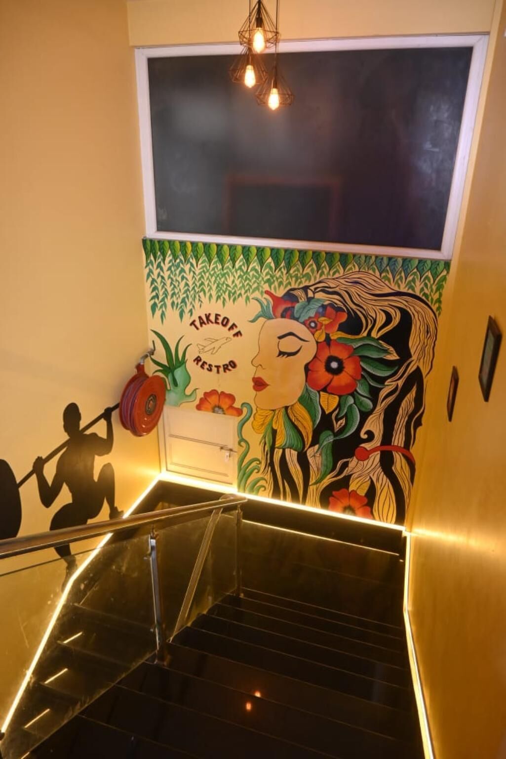 A staircase with a mural featuring a woman and a man lifting weights, a window above, and a ceiling light fixture.
