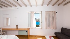 A bedroom with a wooden ceiling, a bed with a white cover, a bedside table with a lamp, a window with curtains, and a view of the sea. - Luxury Sea View Apartment w shared pool (Mykonos)