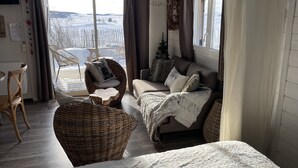 A cozy living room with a sofa, a wicker chair, and a table. There is a view of a snowy landscape through the glass door.