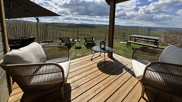 An outdoor patio with wicker furniture, a table, and a bench, overlooking a fenced field and distant hills under a partly cloudy sky.