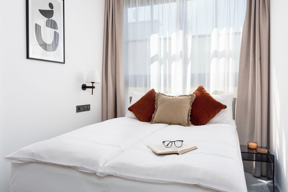 A neatly made bed with white bedding, two pillows, a book, and glasses on the bed.