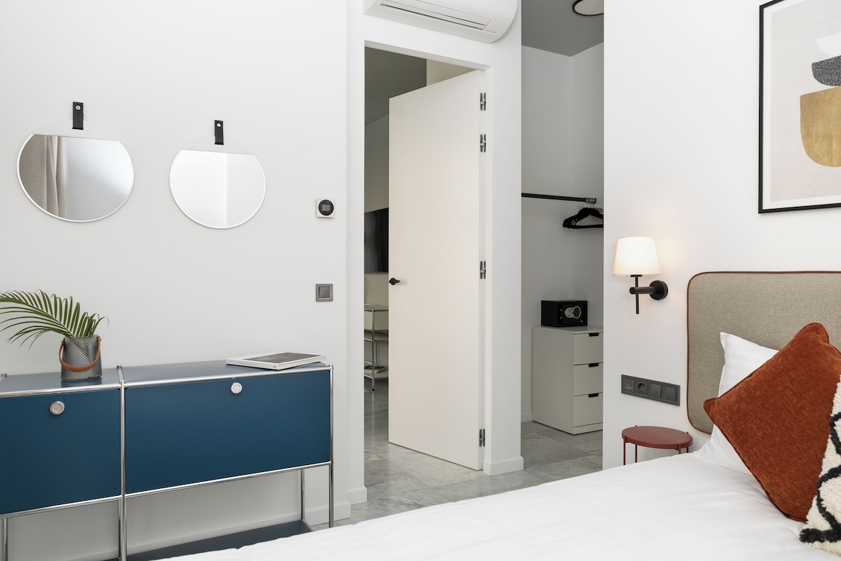 A modern hotel room with a bed, a bedside table, a desk, a mirror, a lamp, and a door leading to another room.