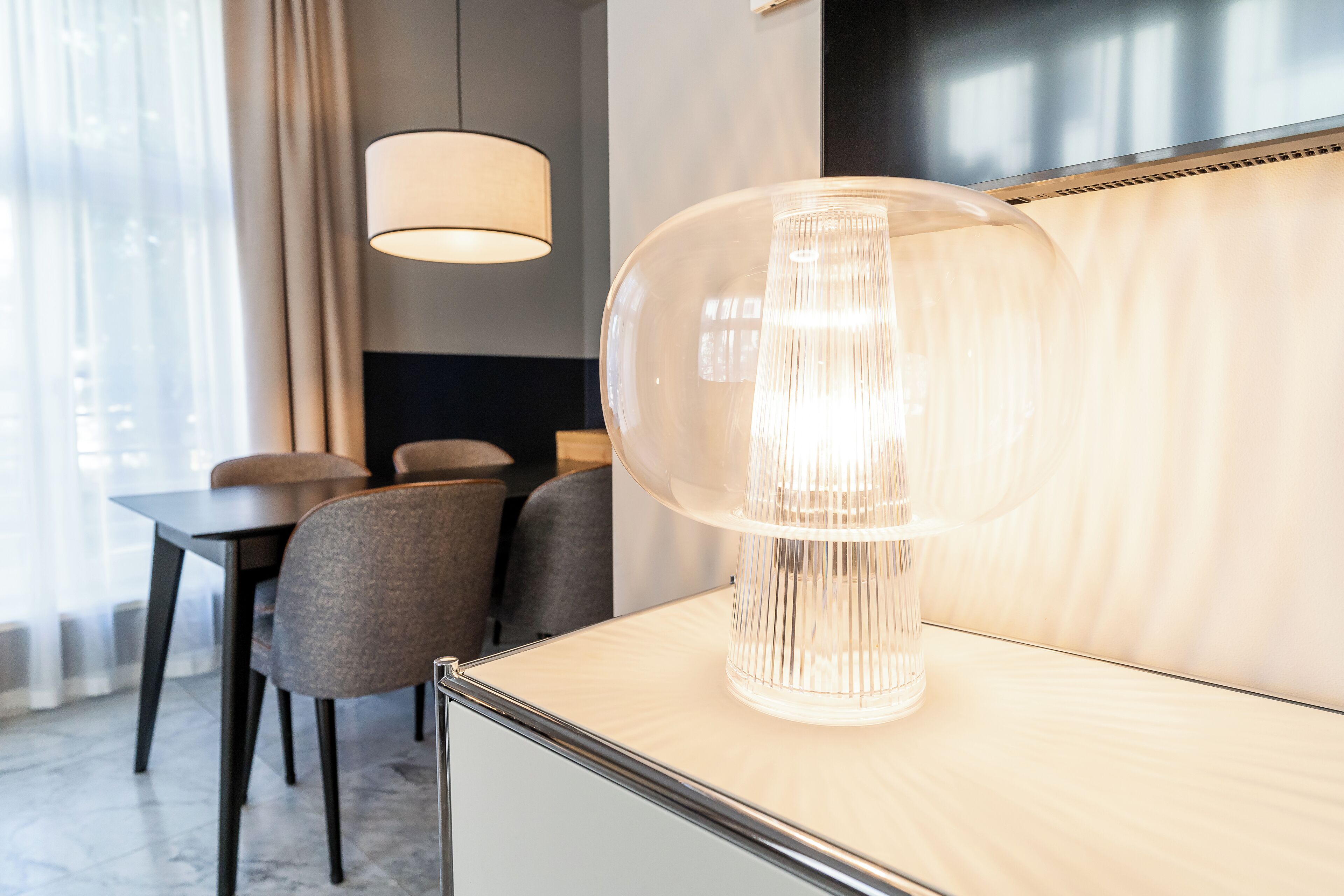 A modern hotel room with a large, clear lamp on a wooden table, a dining area with chairs, and a large window with curtains.
