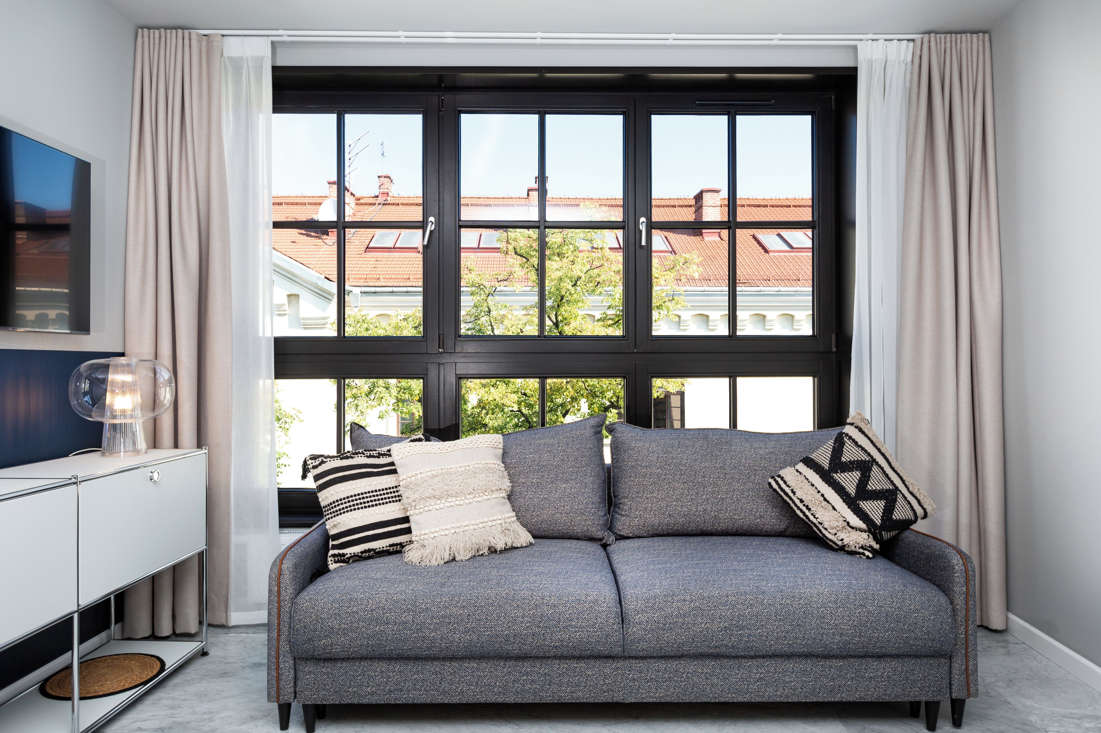 A modern living room with a grey sofa, a TV on the left, and a large window with curtains.