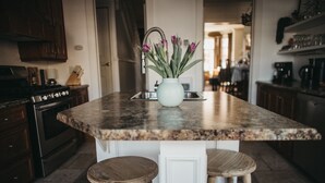 A kitchen island with a white vase of purple flowers, two wooden stools, and a stainless steel sink. - Historic Luxury Downtown Victorian home with Hot Tub, Ocean and Harbour Views (St. John's)