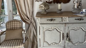 Een kamer met een geruite stoel, een wit kastje met versierde gravures, een lamp en een spiegel met gouden lijst.