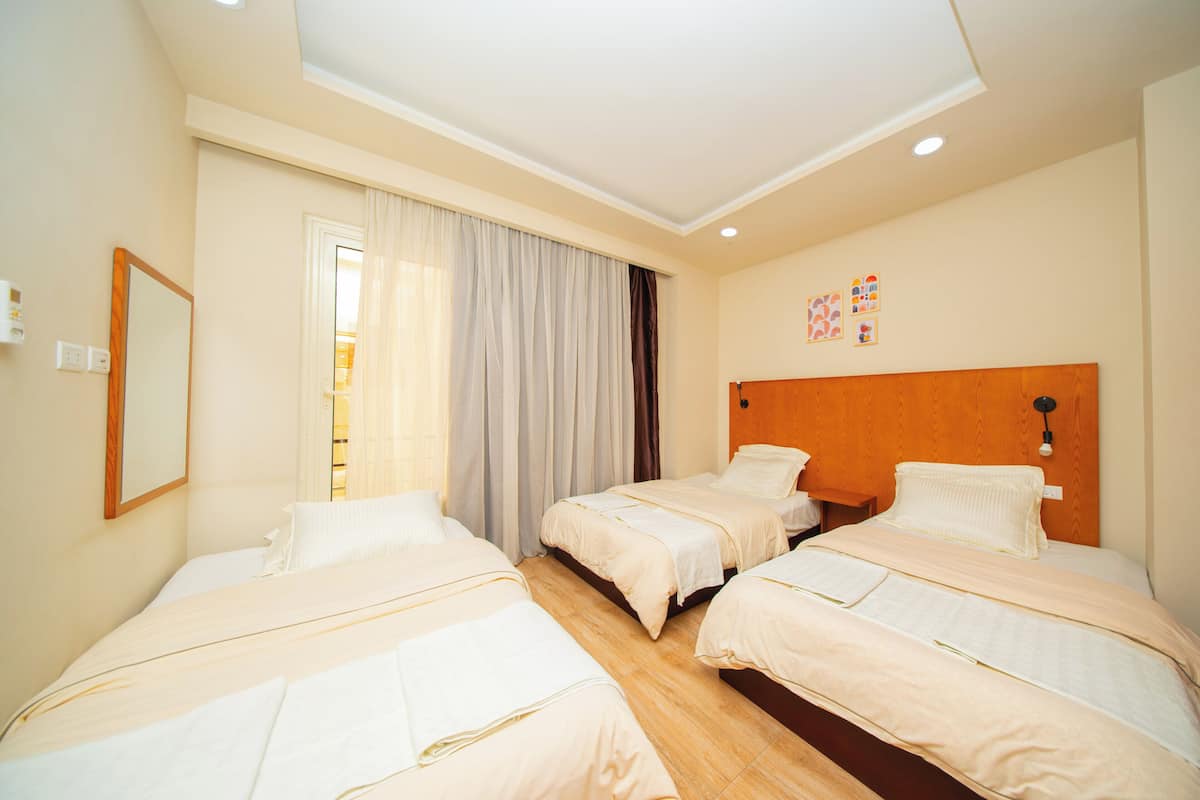 A hotel room with three beds, a wooden headboard, a large window with curtains, and a wall-mounted light fixture.