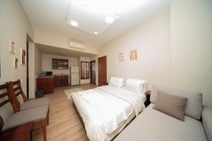 A bedroom with a bed, a sofa, a desk, and a kitchenette in the background.