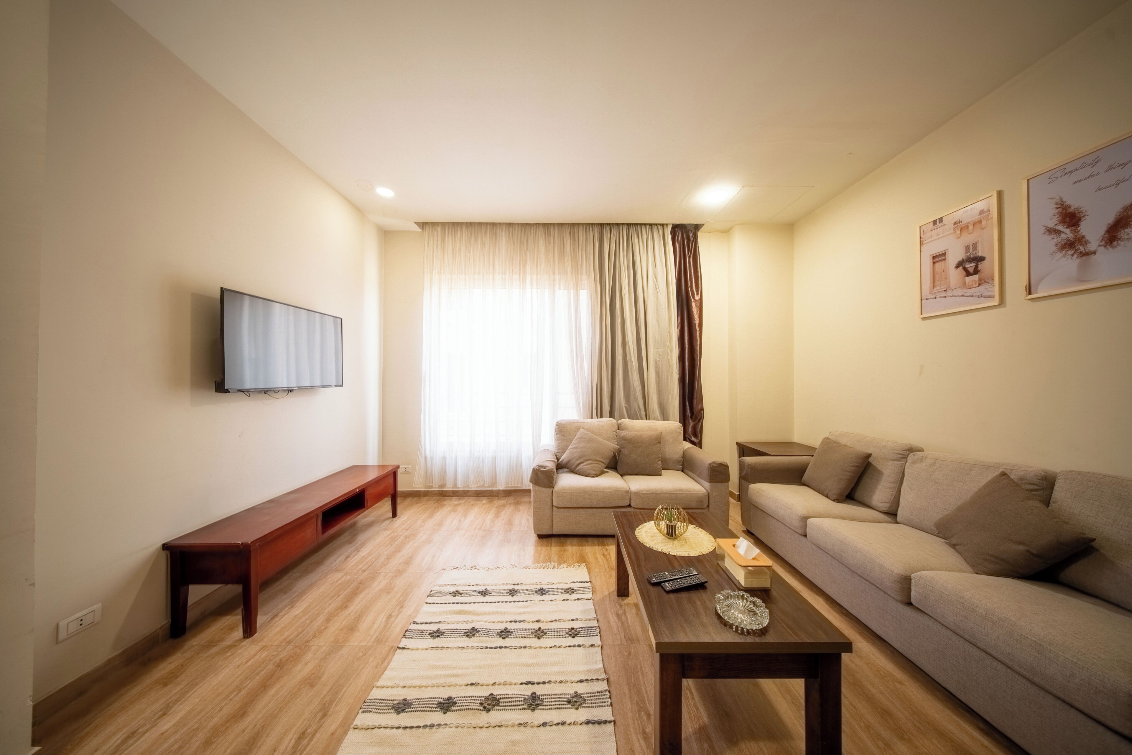 A living room with a sofa, a coffee table, a TV mounted on the wall, and a window with curtains.