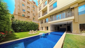 A swimming pool with a clear blue water, surrounded by a well-maintained lawn and a multi-story building with balconies. - The Home (6th of October City)