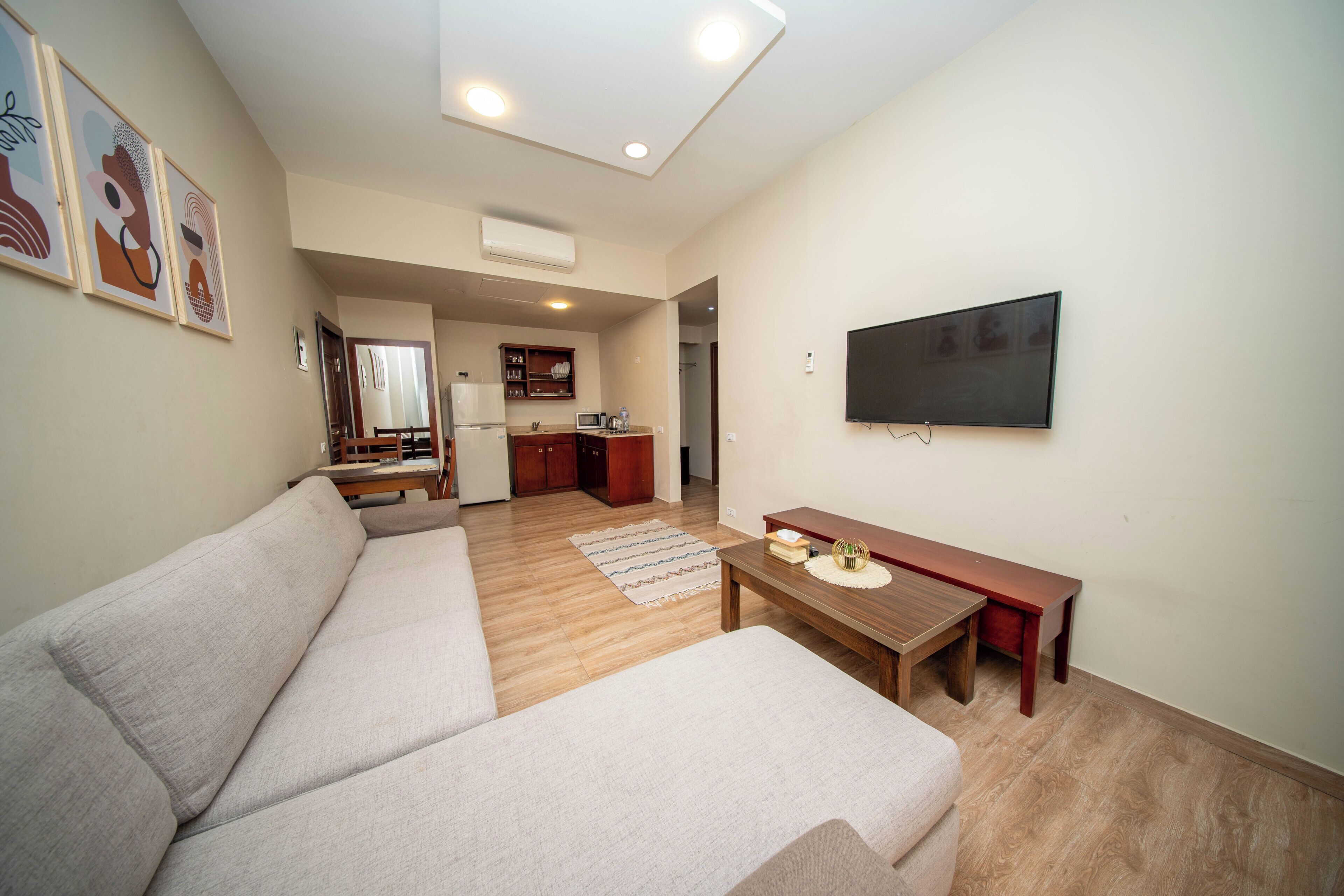 A living room with a sofa, a coffee table, a TV mounted on the wall, and a kitchen area in the background.