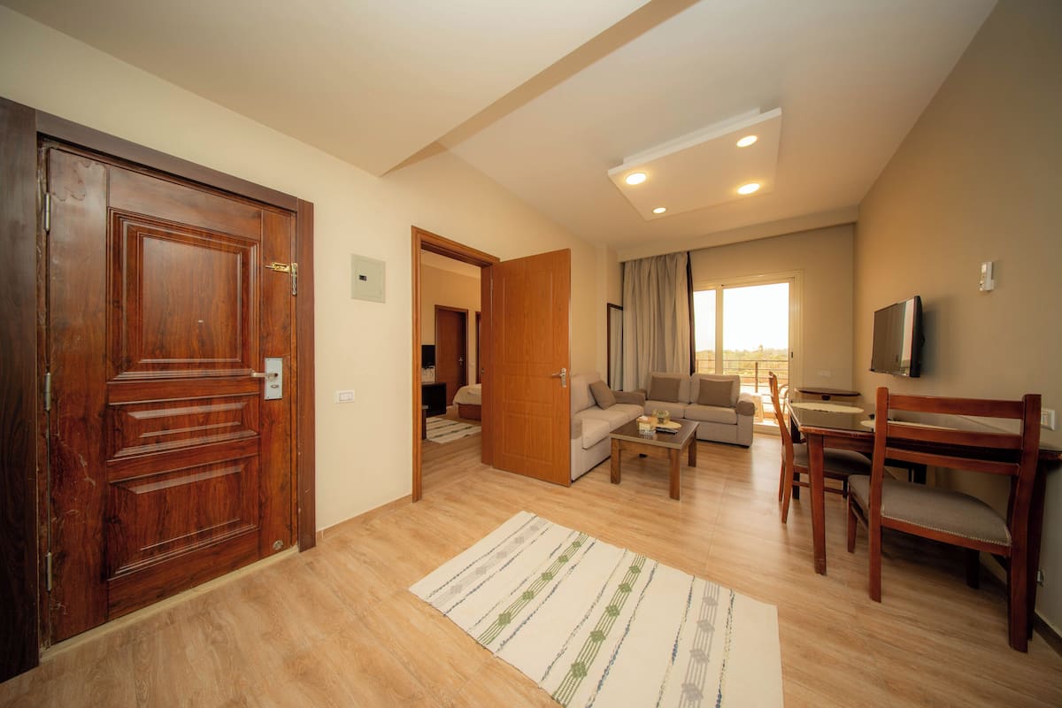 A spacious living area with a wooden door, a sofa set, a dining table with chairs, and a flat-screen TV mounted on the wall.