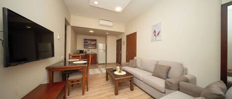 A living room with a sofa, a coffee table, a TV, a dining table with chairs, and a kitchen area in the background.