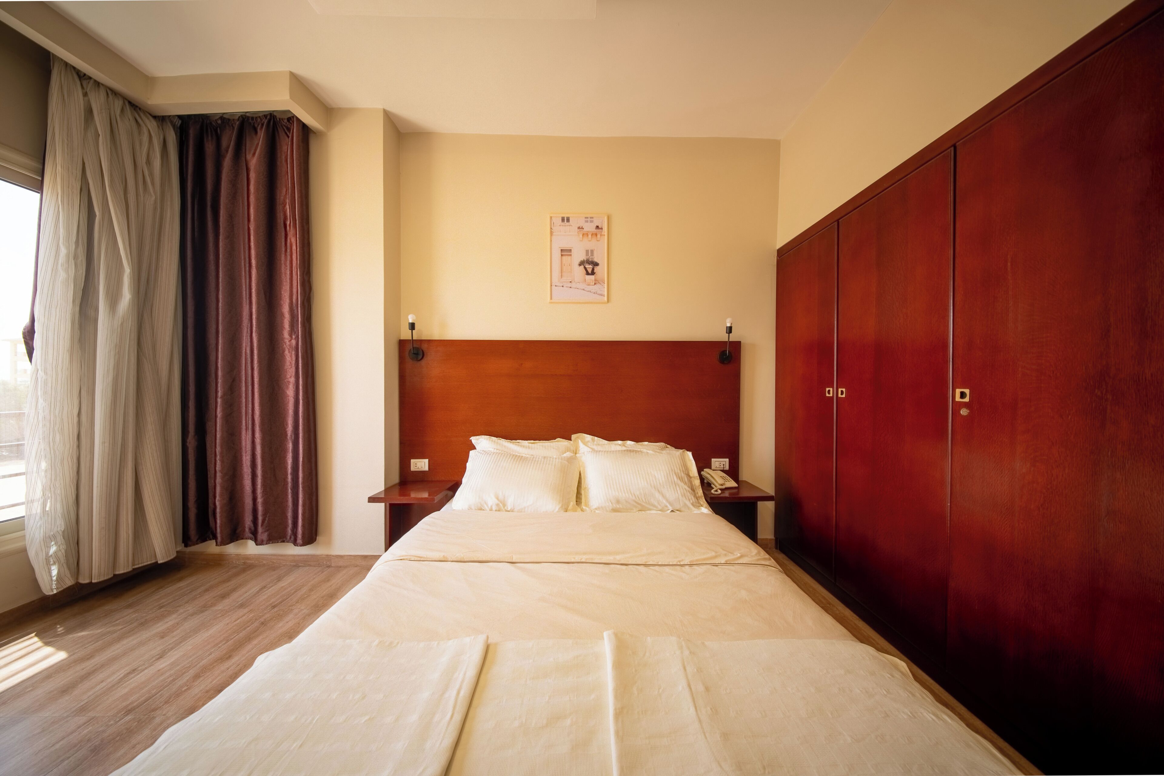 Hotellihuone, jossa on sänky, puinen pääty, vaatekaappi, yöpöytä ja seinälle asennettu puhelin.