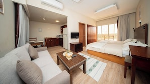 A hotel room with a sofa, a bed, a desk, a TV, and a kitchen area. - The Home (6th of October City)