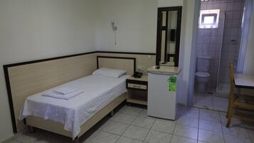 Una habitación pequeña y ordenada con una cama individual, un escritorio pequeño, un espejo, un inodoro y un refrigerador compacto.