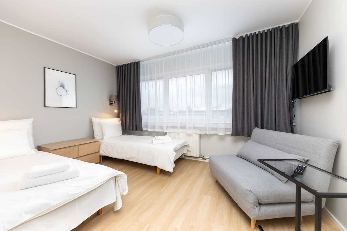 A modern hotel room with two beds, a grey sofa, a flat-screen TV, and a window with sheer curtains.