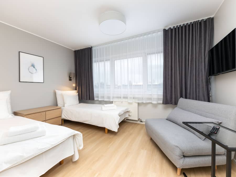 Habitació d'hotel moderna amb dos llits, un sofà gris, televisió de pantalla plana i una finestra amb cortines translúcides.