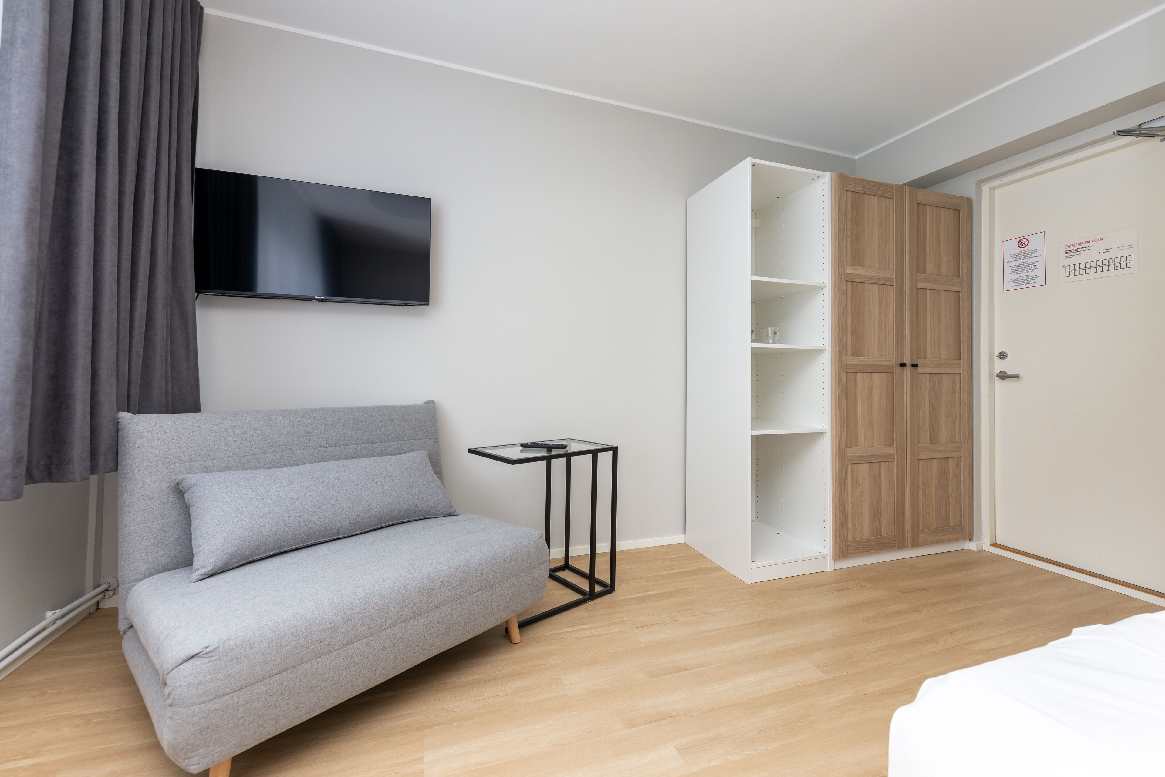 Un dormitorio moderno con un sofá gris, un televisor montado en la pared, un armario de madera y una pequeña mesita de noche negra.