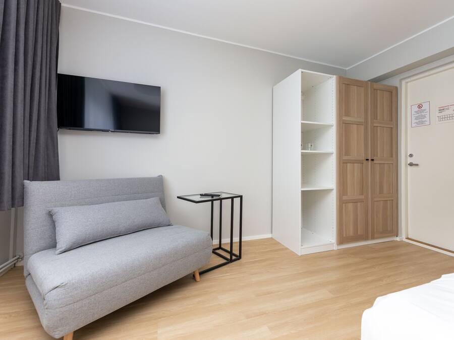 A modern bedroom with a grey sofa, a television mounted on the wall, a wooden wardrobe, and a small black side table.
