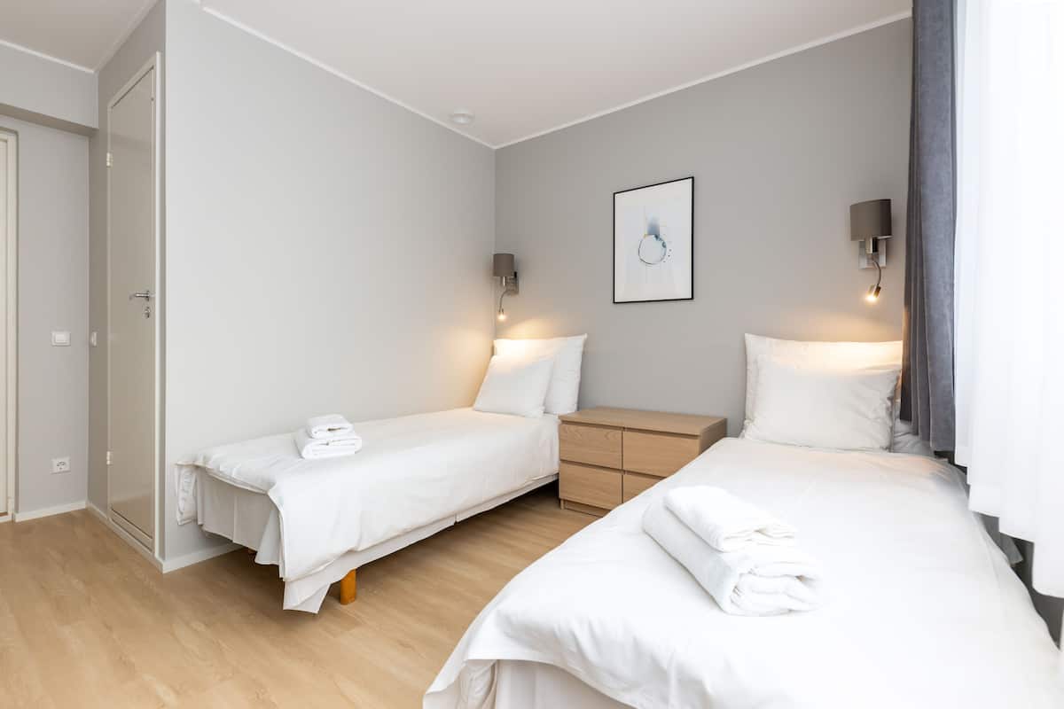 A hotel room with two single beds, a wooden nightstand, a wall-mounted lamp, and a door.
