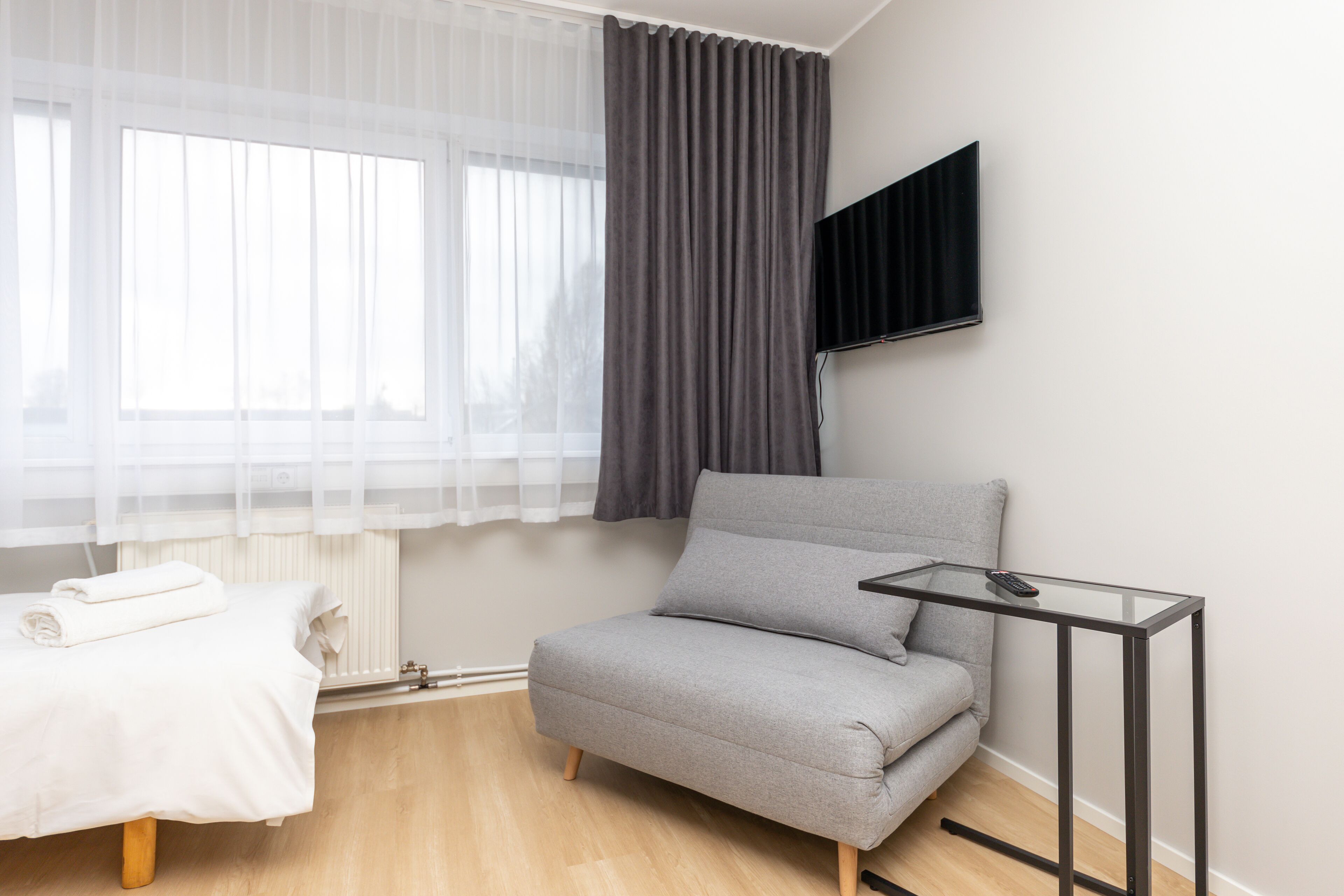 A modern room with a grey sofa, a small glass table, a TV mounted on the wall, and a window with sheer curtains.