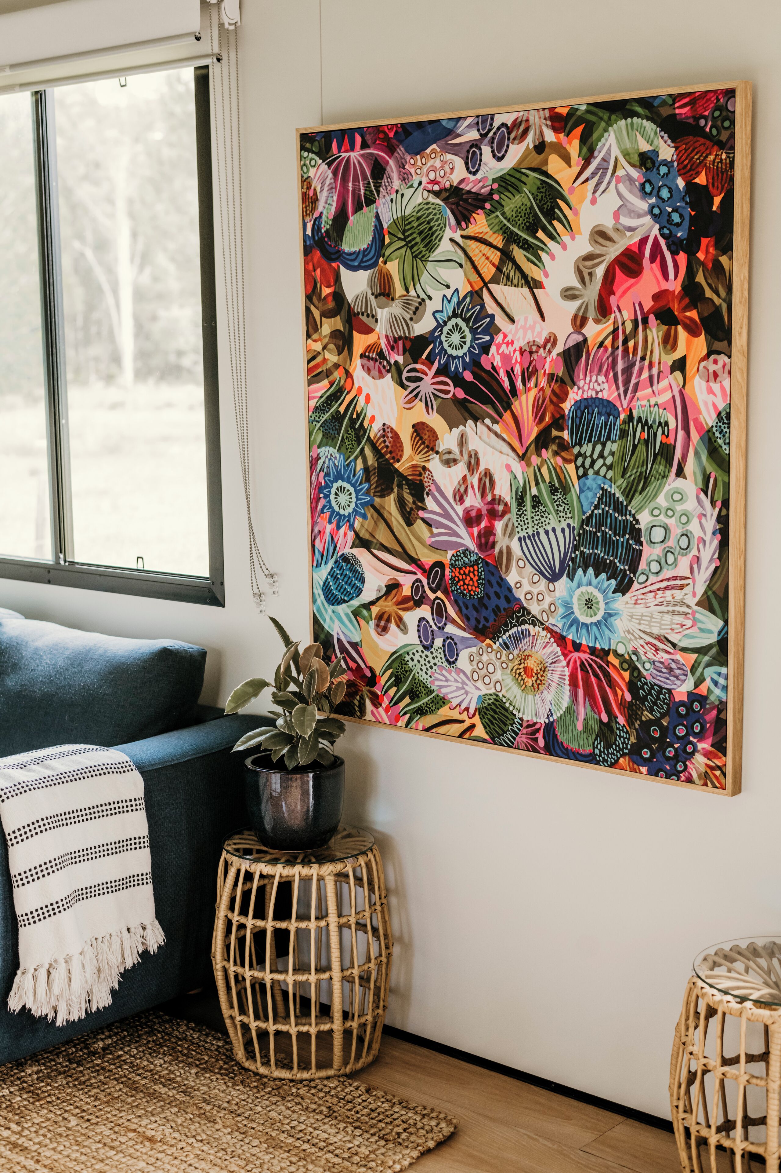Una habitación con un cuadro floral grande en la pared, un sofá azul, una pequeña planta en maceta y una mesita de noche tejida.