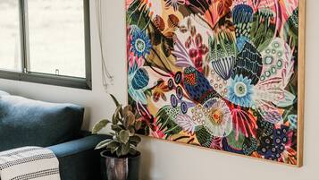 Una habitación con un cuadro floral grande en la pared, un sofá azul, una pequeña planta en maceta y una mesita de noche tejida.