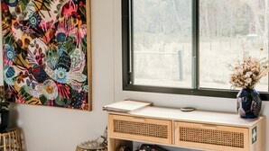 A room with a large window, a colorful floral painting on the wall, a wooden desk with baskets, and a wicker stool. - Contained in Nimbin (Nimbin)