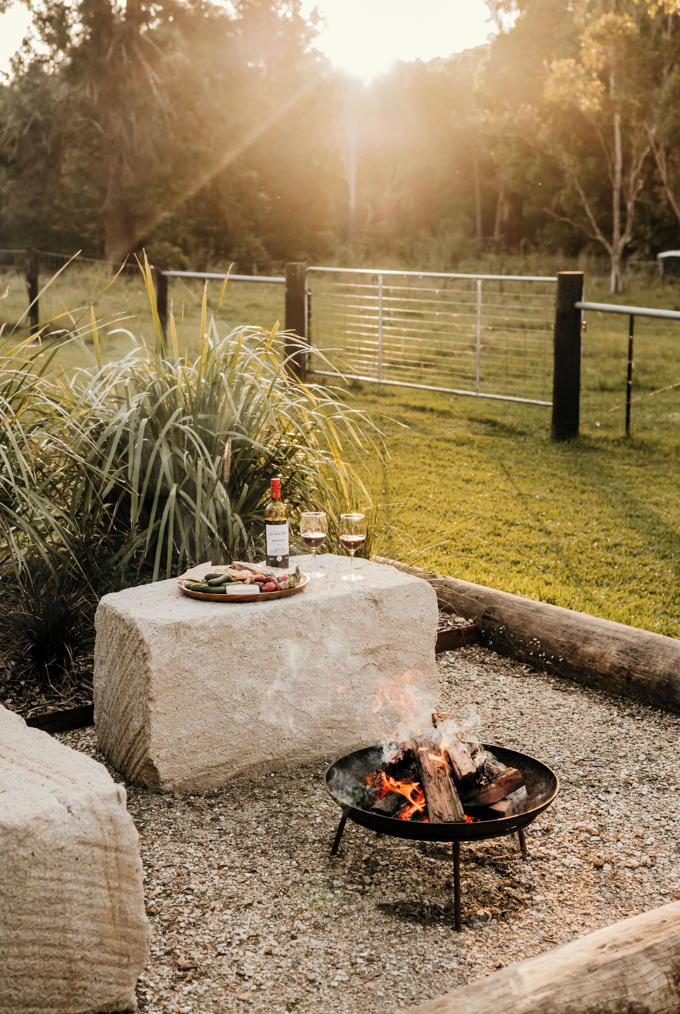 A stone table with a fire pit, surrounded by a gravel area and a grassy field.