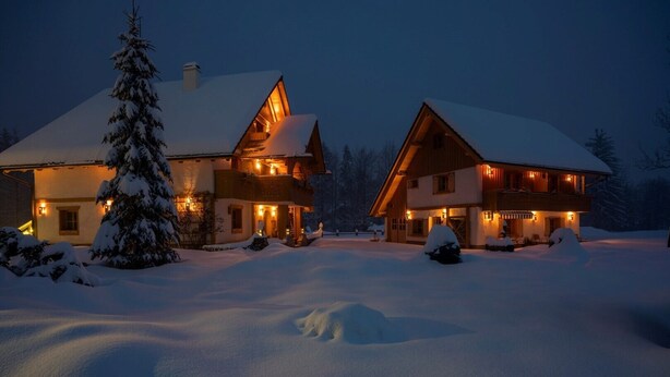 A snow-covered lodge with a large chimney, a snow-covered tree in front, and a well-lit entrance.