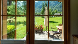 A wooden-framed window with a view of a grassy deck, a table, chairs, and a hanging white cloth.