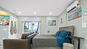Un dormitorio con cama, sofá, reposapiés pequeño y ventana con vista a palmeras.