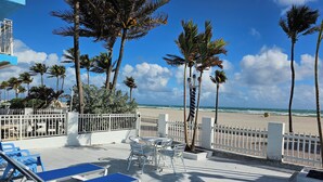 Un patio frente a la playa con sillones y mesas, palmeras y vista al océano.